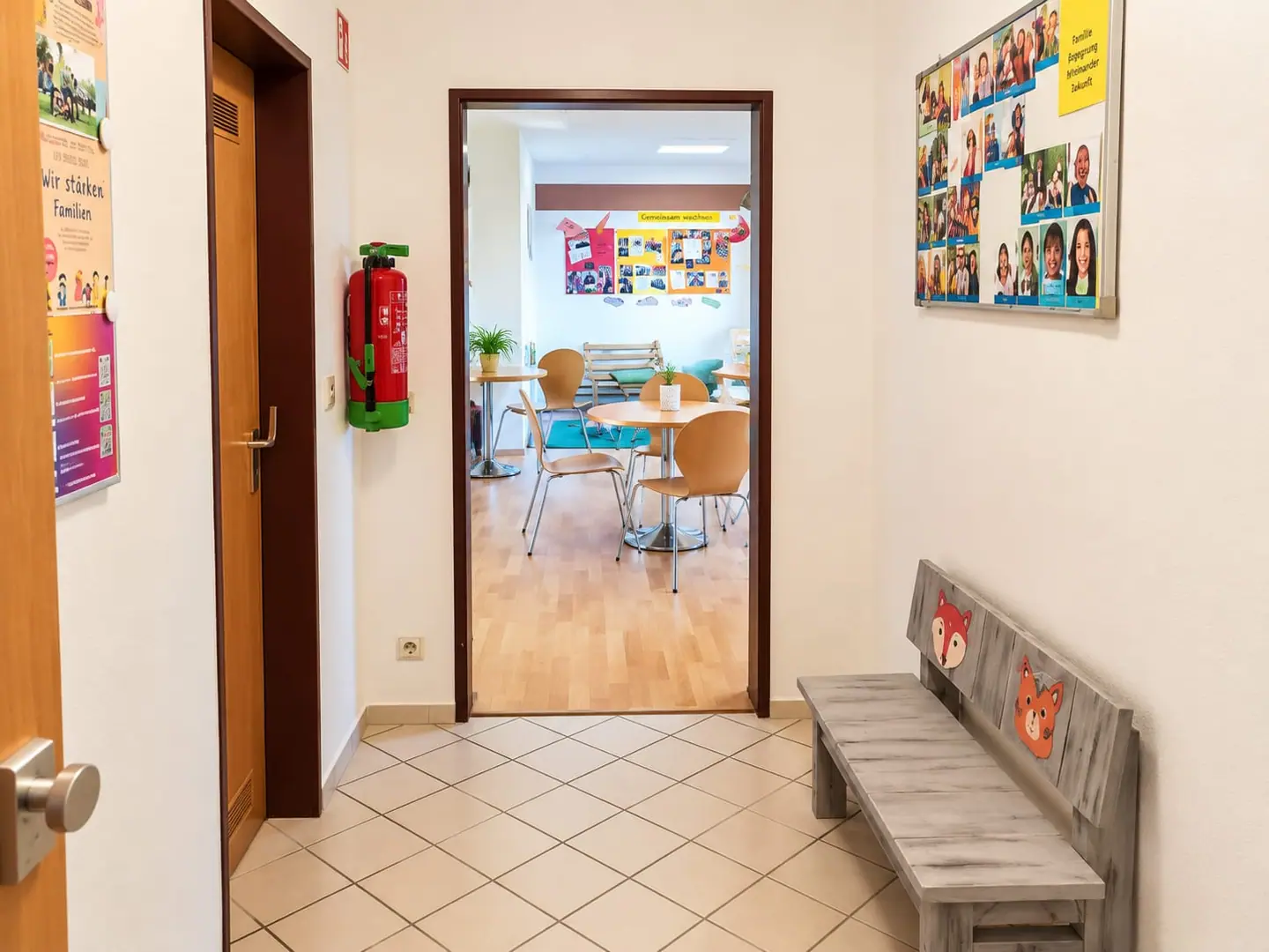 Flur mit Fuchs-Holzbank und Pinnwand voller Fotos; durch die Tür sieht man Tische im Familiencafé.