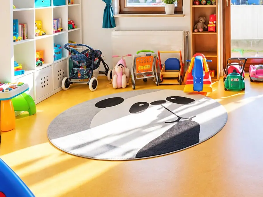 Spielbereich im sonnigen Spielzimmer: runder Panda-Teppich, Lauflernwagen, Schaukelpferd, Puppenwagen und Babyspielzeug.